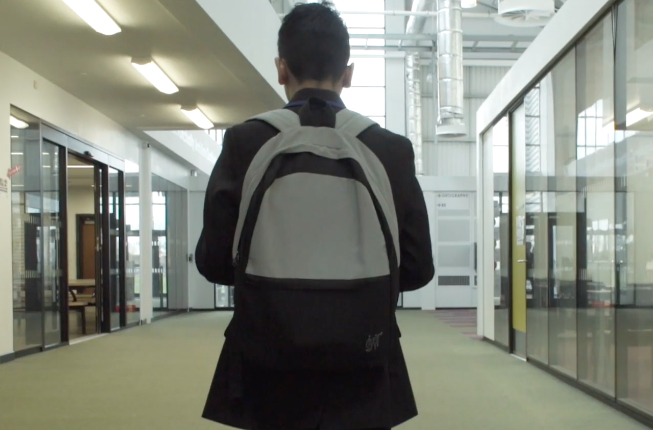 A boy is walking down a school corridor. He is waering a school uniform and has a backpack on.