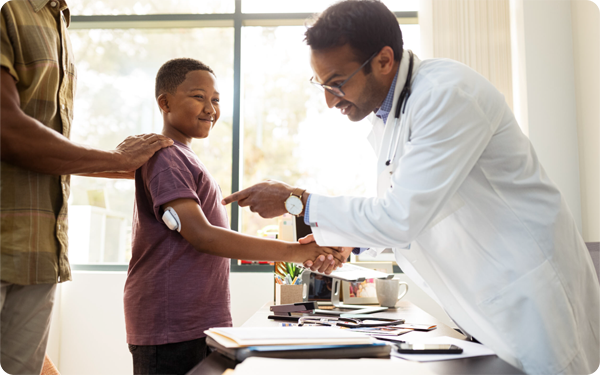 A boy stands with his dad behind him. The boy is shaking a doctors hand. The boy is wearing an omnipod.