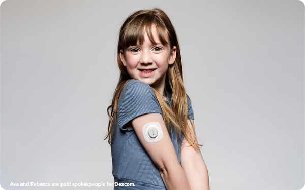 A little girl smiles and looks at the camera, she has a glucose monitor on her arm.