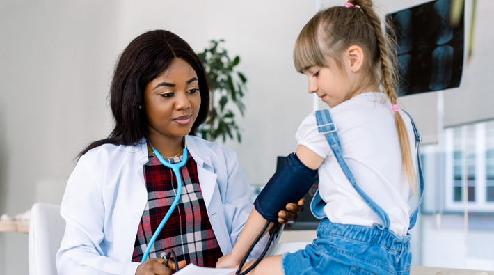A little girl sits with a doctor. The doctor is checking her blood pressure.